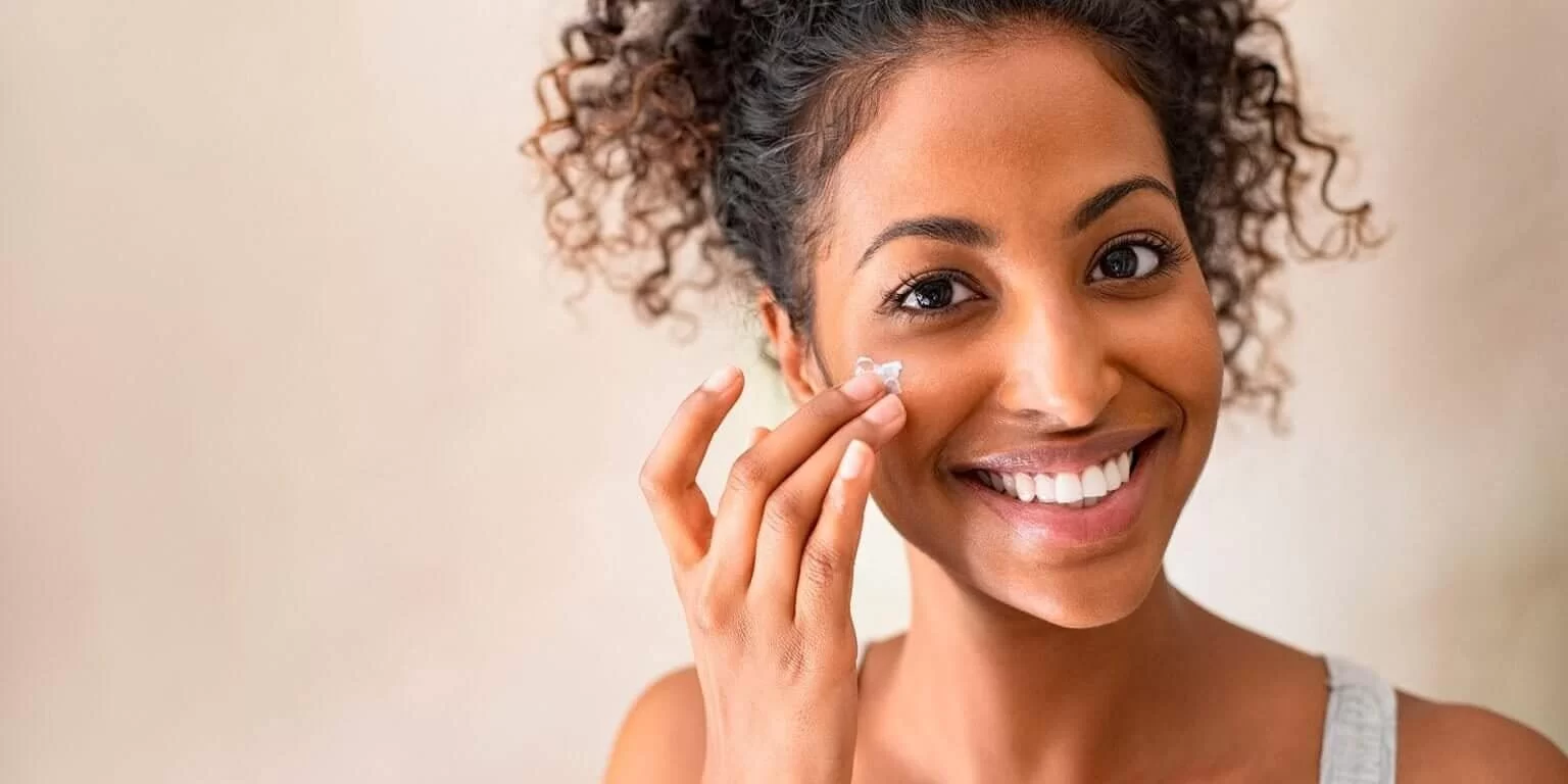 Mulher jovem de ascendência africana com cabelo cacheado preso em coque, sorrindo e aplicando creme facial na bochecha, com fundo bege claro, destacando uma rotina de cuidados com a pele no outono.